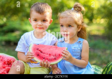 Zwei Kinder sitzen zwischen Wassermelonen im Garten. Kinder essen Obst im Freien. Gesunde Snack für Kinder. 2 Jahre altes Mädchen und Junge genießen Wassermelone Stockfoto