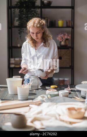Weibliche Töpfer sitzen und rührt malen mit einem Pinsel eine Tasse auf den Tisch. Frau, die Keramik-Element. Töpferarbeiten, handgemachte und kreative Fähigkeiten. Stockfoto