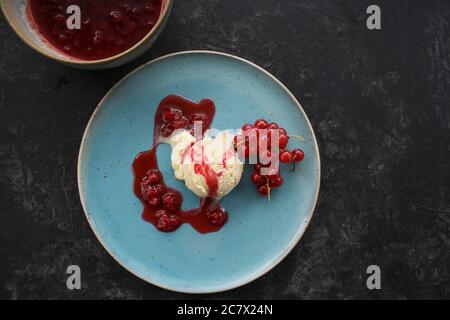 Vanilleeis mit roten Johannisbeeren und Fruchtgeleesauce auf blauem Teller, dunkler Hintergrund mit Kopierraum, Hochwinkelansicht von oben, ausgewählter Fokus Stockfoto