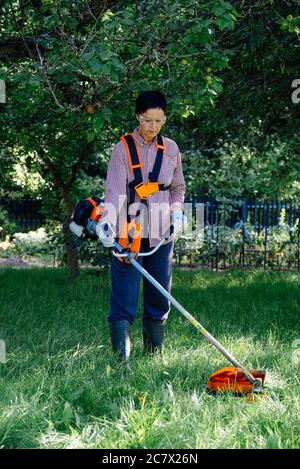 Erwachsene Frau mäht das Gras im Hinterhof mit String Trimmer. Gartenarbeit Konzept. Stockfoto