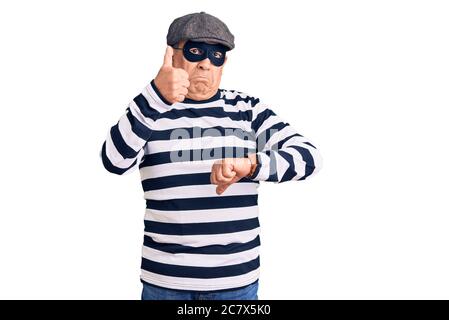 Senior stattlichen Mann trägt Einbrecher Maske und T-Shirt tun Daumen nach oben und unten, Meinungsverschiedenheiten und Vereinbarung Ausdruck. Verrückte Konflikt Stockfoto