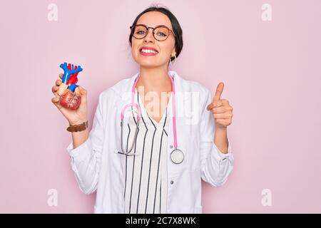 Junge schöne Kardiologin Frau mit blauen Augen trägt Stethoskop halten Herz glücklich mit großen Lächeln tun ok Zeichen, Daumen mit Fingern, excelle Stockfoto