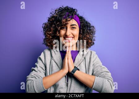 Schöne lockige arabische Sportlerin Sport treiben tragen Sportkleidung über lila Hintergrund beten mit Händen zusammen um Vergebung bitten lächelnd conf Stockfoto