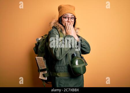 Junge Wanderfrau trägt Wanderrucksack, Kantine und Karte über gelbem Hintergrund schockiert über Mund mit Händen für Fehler. Geheimkonzept. Stockfoto