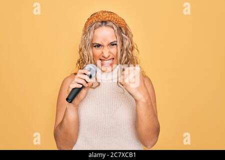 Junge schöne blonde Sängerin Frau singen mit Mikrofon über gelben Hintergrund verärgert und frustriert schreien vor Wut, schreien verrückt mit ang Stockfoto