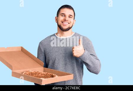 Junge gut aussehende Mann hält Lieferung italienische Pizza lächelnd glücklich und positiv, Daumen nach oben tun ausgezeichnet und Genehmigung Zeichen Stockfoto