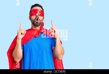 Junger hispanischer Mann mit Superhelden Kostüm erstaunt und überrascht, nach oben schauen und zeigen mit Fingern und erhobenen Armen. Stockfoto