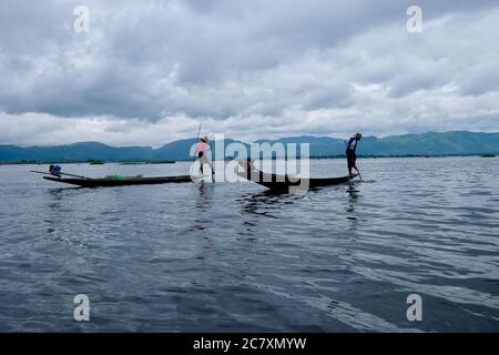Zwei Fischer stehen auf einem kleinen Boot im welligen Inle See. Mit Bein zu rudern Boot. Weißer wolkig Himmel mit Bergen Stockfoto