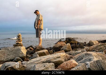 Ein Mann, der neben einem Steinkairn an der Küste des Ozeans in Connecticut, USA, in der Nähe des Harkness Memorial State Park steht Stockfoto