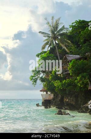 Grüner Kokosnussbaum und Blockhütte auf Klippe über dem grünen Ozean. Herrlicher weißer, wolkiger, sonniger Himmel. Eine Ecke der Insel Boracay auf den Philippinen. Stockfoto
