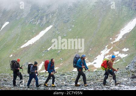 Wanderteam zu Fuß auf felsigen Weg mit hohen Berghang im Hintergrund. Männliche Touristen mit Rucksäcken und Wanderstöcken wandern in der bergigen Region. Konzept des Reisens, Tourismus, aktive Freizeit. Stockfoto