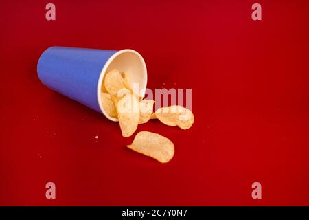 Nahaufnahme von Kartoffelchips, die aus einem verschüttet werden Blauer Papierbecher auf rotem Hintergrund Stockfoto