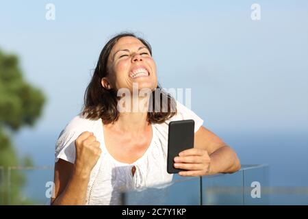 Aufgeregt Erwachsene Frau feiert Nachrichten auf Smartphone auf einem Balkon am Strand Stockfoto