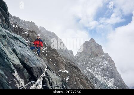 Junger Mann in Sonnenbrille und Schutzhelm hält festsitzend Seil beim Klettern Berg. Alpinistische, aufsteigende natürliche Felsformation. Konzept des Bergsteigens und des alpinen Kletterns. Stockfoto