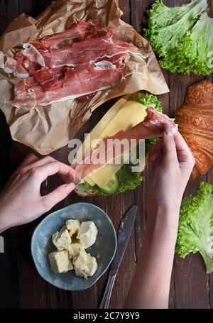 Hände halten Marmelade Fleisch Scheiben und rustikalen Holztisch mit Sandwich Croissant, grünen Salatblättern, frische Gurken, Käse, Butter und Messer, flach Stockfoto