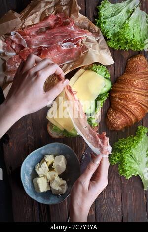 Hände halten Marmelade Fleisch Scheiben und rustikalen Holztisch mit Sandwich Croissant, grünen Salatblättern, frische Gurken, Käse, Butter und Messer, flach Stockfoto