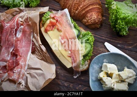 Croissant-Sandwich mit Marmelade-Scheiben, grünen Salatblättern, frischen Gurken, Käse, Butter und Messer, auf rustikalem Holztisch, flatla Stockfoto