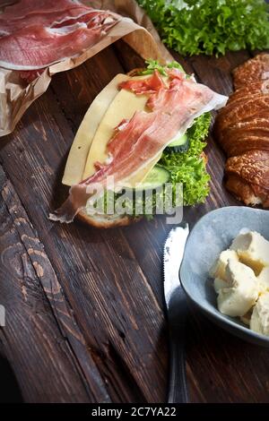 Croissant-Sandwich mit Marmelade-Scheiben, grünen Salatblättern, frischen Gurken, Käse, Butter und Messer, auf rustikalem Holztisch, flatla Stockfoto