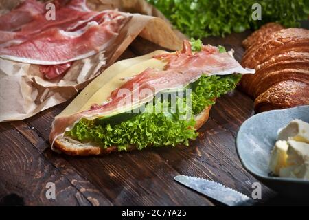 Croissant-Sandwich mit Marmelade-Scheiben, grünen Salatblättern, frischen Gurken, Käse, Butter und Messer, auf rustikalem Holztisch, flatla Stockfoto