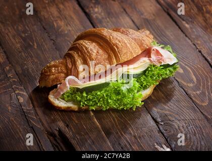 Ein Croissant-Sandwich mit Jamon-Fleischscheiben, grünen Salatblättern, frischen Gurken und Käse, auf rustikaler Holztischoberfläche, Copyspace Stockfoto