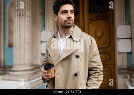 Junger attraktiver Mann im Trenchcoat mit Kaffee, um träumerisch weg zu gehen, indem er durch die Straße geht Stockfoto