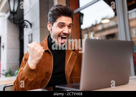 Junger, fröhlicher, zwangloser Mann, der freudig mit Ja-Geste arbeitet, während er im Café auf der Straße am Laptop arbeitet Stockfoto