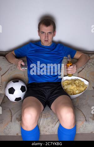 Junger gelangweilter Mann in Fußballuniform sitzt im Wohnzimmer und schaut mit Chips und Bier fernzusehen Stockfoto