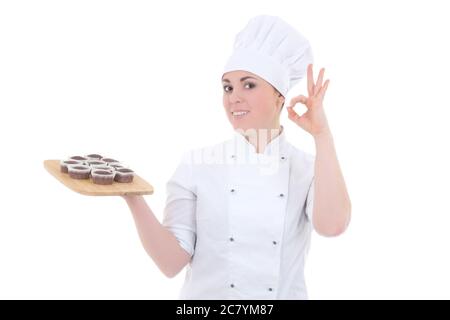 Junge attraktive Köchin Frau in Uniform mit Schokolade Muffins zeigt ok Zeichen isoliert auf weißem Hintergrund Stockfoto