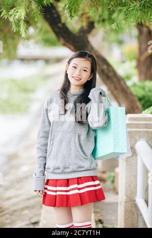 Portrait von schönen positiven Teenager asiatische Mädchen in hellgrau Hoodie mit Einkaufstasche und Blick auf die Kamera Stockfoto