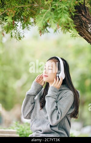 Schöne junge vietnamesische Teenager-Mädchen lächeln und Augen schließen, wenn sie ihre Lieblingsmusik in Kopfhörer hören Stockfoto