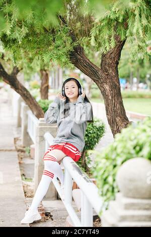 Happy Teenage asiatische Mädchen auf Geländer im Park sitzen und Musik in Kopfhörern hören Stockfoto