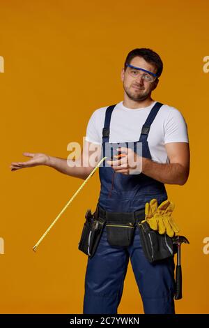 Verwirrt kaukasischen Werkstatt Arbeiter in Uniform halten Maßband und zuckende Schultern. Handwerker trägt Werkzeug Gürtel isoliert auf gelben Studio-Hintergrund Stockfoto