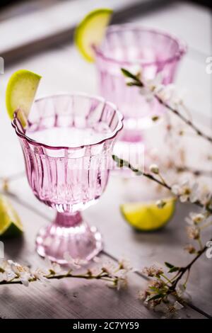 2 Gläser harter Seltzer in vintage rosaroten Gläsern mit einer Limettengarnitur serviert. Gläser werden auf rustikalem weißen Holztisch mit Apfelblüte gesetzt Stockfoto