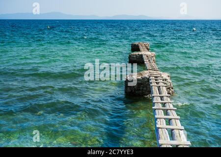 Ein schmaler Steg aus zwei Holzplanken führt über mehrere Poller ins türkisblaue Mittelmeer in Griechenland und lädt zum Baden und Baden ein Stockfoto