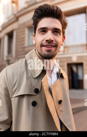 Junge attraktive stilvolle Mann in Trenchcoat glücklich Blick weg im Freien Stockfoto