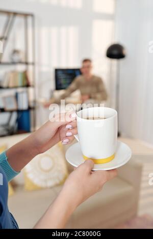 Hände der Frau, die Tasse Kaffee zu ihrem Mann, der am Computer zu Hause wegen Pandemie arbeitet Stockfoto