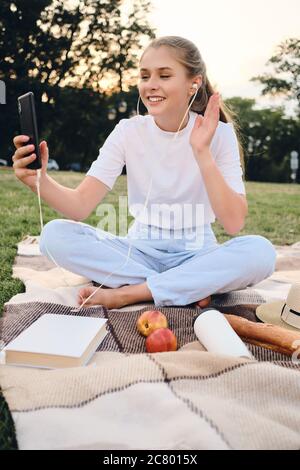 Ziemlich lächelndes Teenager-Mädchen, das auf Karos mit Ohrhörern sitzt und sich im Handy beim Picknick im Park fröhlich im Videochat unterhielt Stockfoto