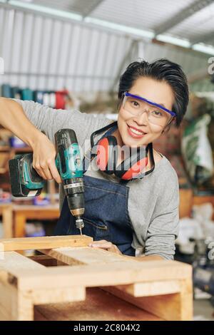 Portrait von glücklichen Zimmermann in Schutzbrille mit Bohrer bei der Herstellung von Holzmöbeln in der Werkstatt Stockfoto