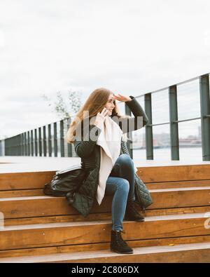 Schöne Frau mit langen Haaren am Telefon reden und auf der Suche nach etwas in der Ferne. Herbst oder Winter, Mädchen in Outdoor-Jacke auf Holz sitzen Stockfoto