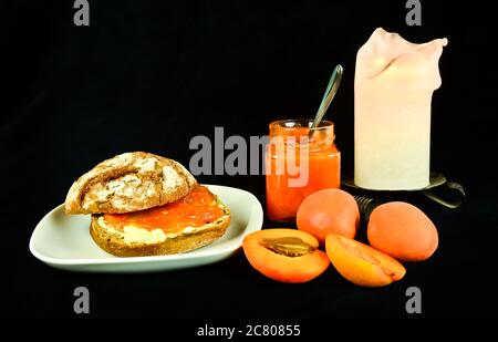 Ein Stillleben mit Aprikosen, Aprikosenmarmelade und einem Brötchen mit Butter und Marmelade, bereit zum Essen vor schwarzem Hintergrund Stockfoto