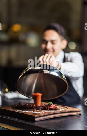 Glückliche junge Kellner im Smoking und Fliege anheben Metall Cloche aus Serviertablett Stockfoto