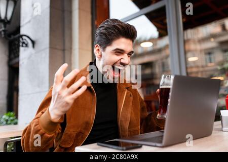 Junger, positiv emotionaler Mann, der freudig schreit, während er im Café auf der Straße am Laptop arbeitet Stockfoto