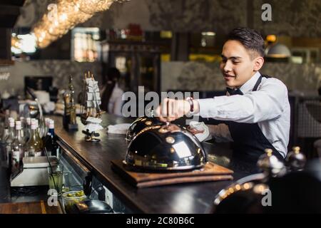 Attraktive junge Kellner im Smoking hält Serviertablett mit Metall Cloche und Serviette Stockfoto