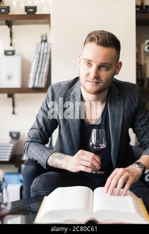 Sommelier untersucht Wein. Zugeschnittenes Bild eines selbstbewussten männlichen Sommeliers, der Wein untersucht, während er ihn riecht und sich an den Holztisch mit Weinregal i lehnt Stockfoto