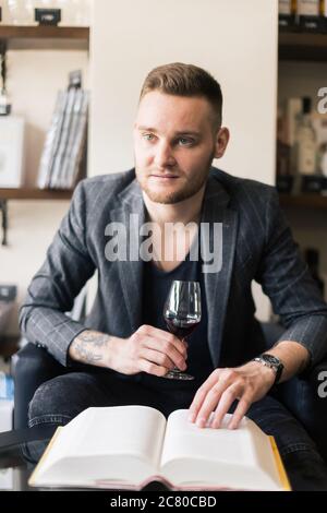 Sommelier untersucht Wein. Zugeschnittenes Bild eines selbstbewussten männlichen Sommeliers, der Wein untersucht, während er ihn riecht und sich an den Holztisch mit Weinregal i lehnt Stockfoto