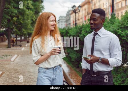Schwarzer Mann und rothaarige Unternehmerinnen, die draußen auf der Straße spazieren und während der Pause Kaffee trinken. Unternehmer reden und diskutieren Projekt Stockfoto