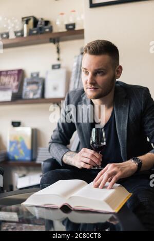 Sommelier untersucht Wein. Zugeschnittenes Bild eines selbstbewussten männlichen Sommeliers, der Wein untersucht, während er ihn riecht und sich an den Holztisch mit Weinregal i lehnt Stockfoto