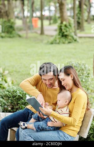 Glückliche junge Eltern verbringen Zeit mit ihrem kleinen Sohn im Park, sitzen auf der Bank und zeigen interessante Buch zu Kind Stockfoto