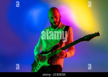 Gespannt. Junge kaukasische inspirierte und expressive Musiker, Gitarrist auf bunten Hintergrund in Neon. Konzept der Musik, Hobby, Festival, Kunst. Fröhliche Künstler, bunt, helles Porträt. Stockfoto