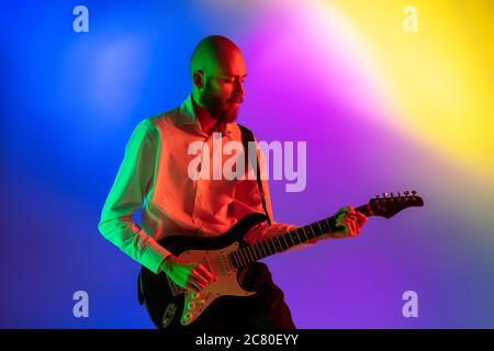 Gespannt. Junge kaukasische inspirierte und expressive Musiker, Gitarrist auf bunten Hintergrund in Neon. Konzept der Musik, Hobby, Festival, Kunst. Fröhliche Künstler, bunt, helles Porträt. Stockfoto